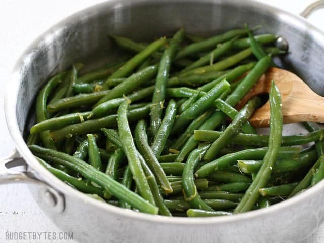 Side view of a skillet full of Lemon Butter Green Beans with a wooden spoon