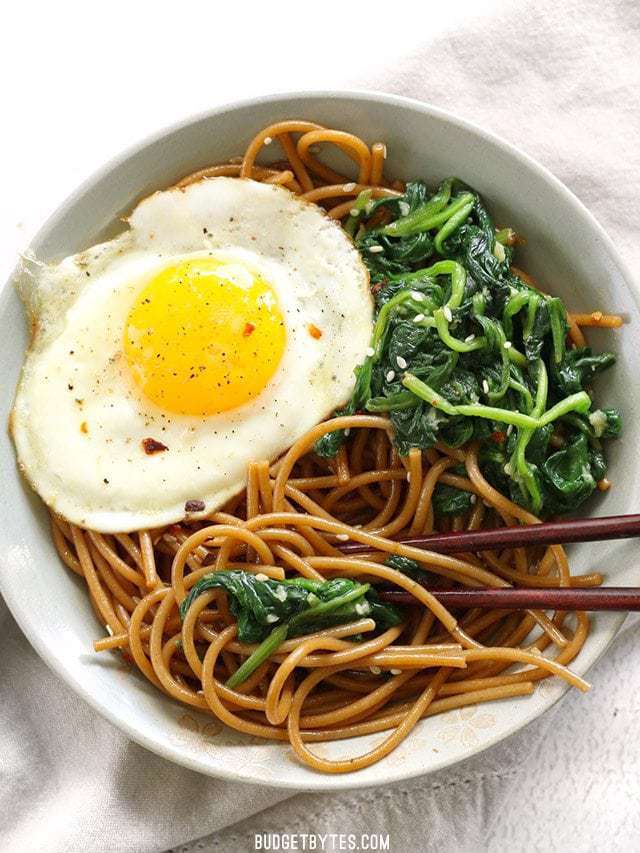 A bowl of Sesame Noodles with Wilted Greens and a fried egg with chopsticks, viewed from above