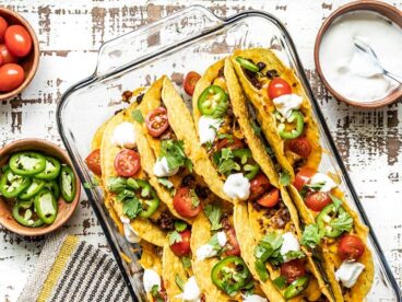 A glass casserole dish filled with baked beef and black bean tacos, with toppings in bowls on the sides