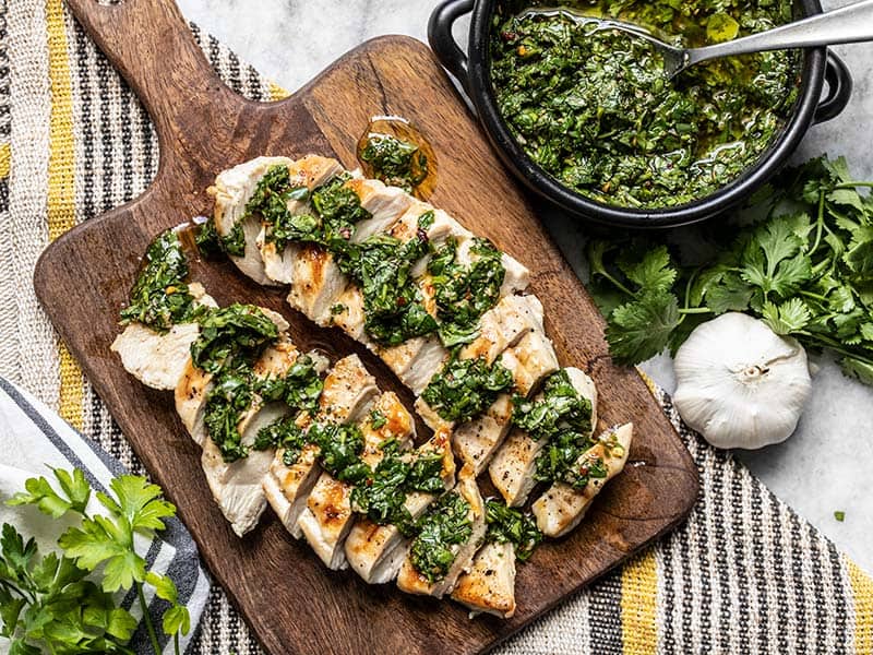 Sliced chicken breast on a wooden cutting board, drizzled with chimichurri sauce. A bowl of chimichurri on the side.