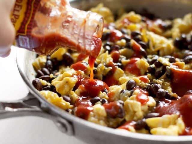 Taco sauce being poured onto the eggs and beans in the skillet