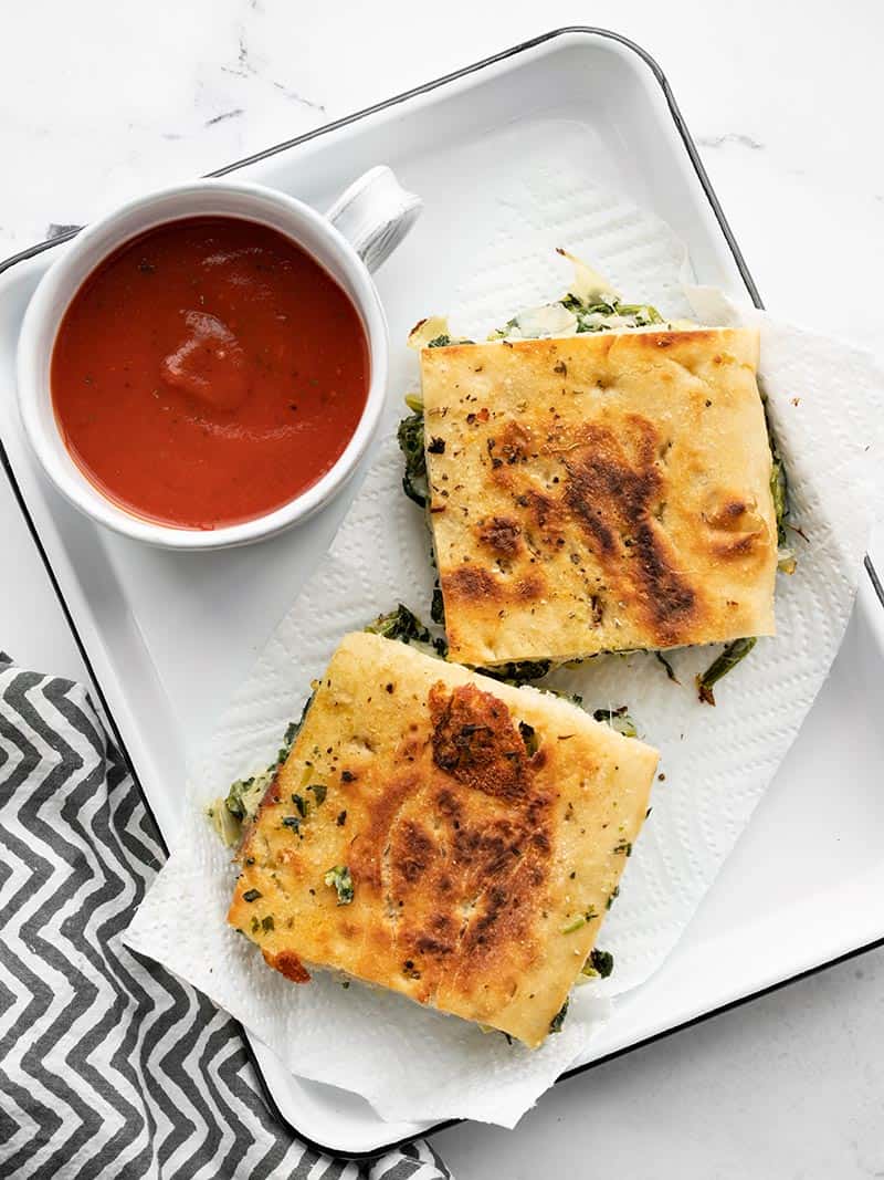 Two spinach artichoke grilled cheese sandwiches on a serving tray with a cup of tomato soup