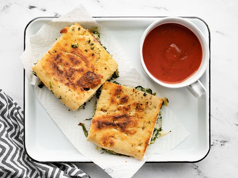 Two spinach artichoke grilled cheese sandwiches on a serving tray with a cup of tomato soup.