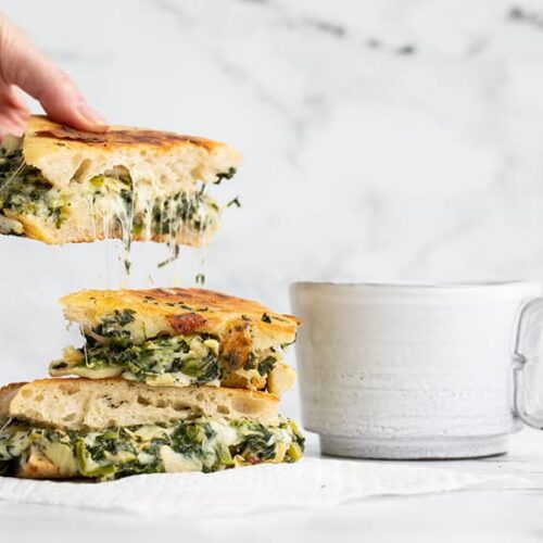 A hand picking up a piece of spinach artichoke grilled cheese with cheese stretching from the slice