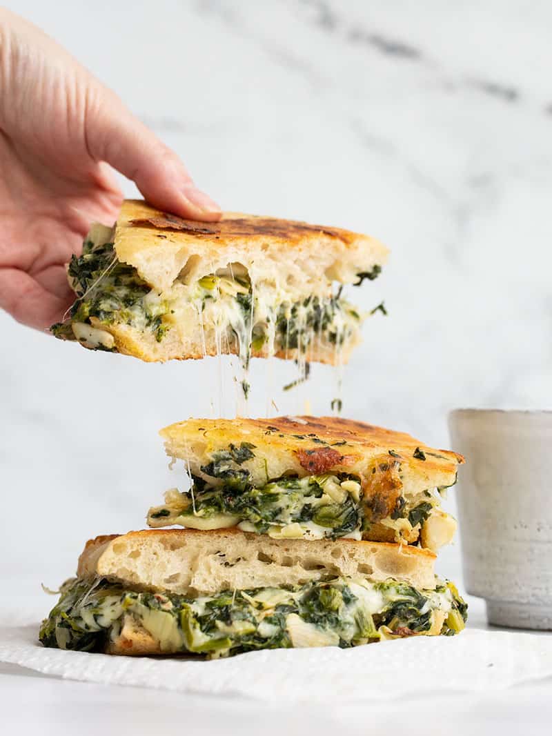 A hand picking up half of a spinach artichoke grilled cheese with cheese stretching between the pieces