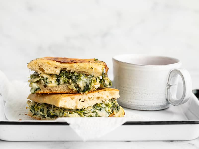 Two spinach artichoke grilled cheese sandwiches stacked on a serving tray next to a mug of tomato soup