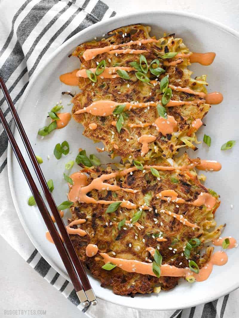 Two Savory Cabbage Pancakes on a plate drizzled with sriracha Mayo, green onion, and sesame seeds.