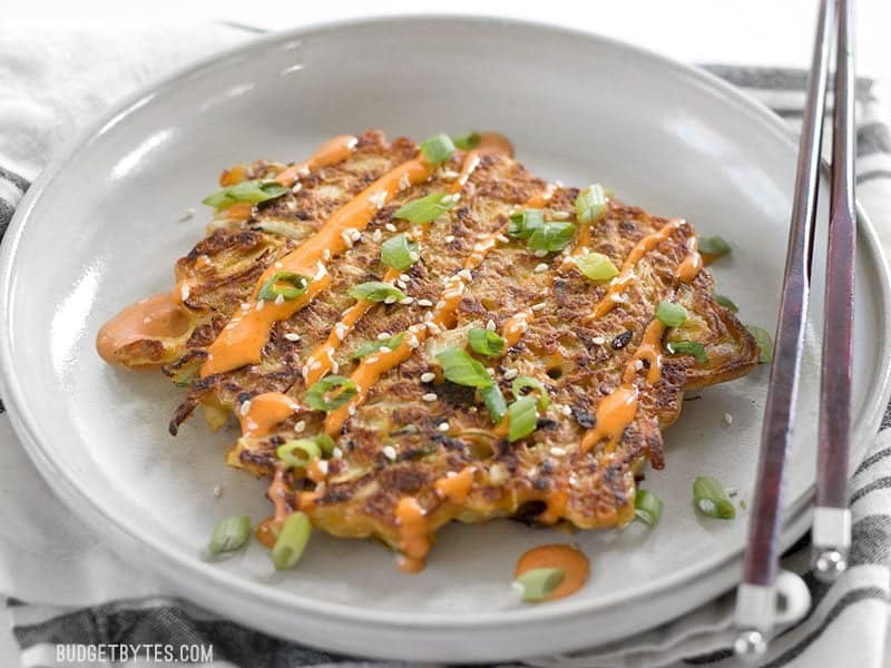 Front view of one Savory Cabbage Pancake on a plate with chopsticks, sriracha mayo, green onion, and sesame seeds