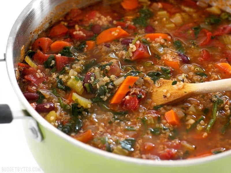 Close up side view of the pot of Garden Vegetable Quinoa Soup with a wooden spoon lifting some of the vegetables