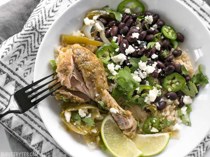 A bowl of Slow Cooker Salsa Verde Chicken with rice, beans, queso fresco, and cilantro. A fork pulling the meat from the bone.