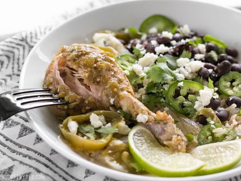 A fork pulling the meat from a Slow Cooker Salsa Verde Chicken drumstick in a bowl with rice, beans, and cheese