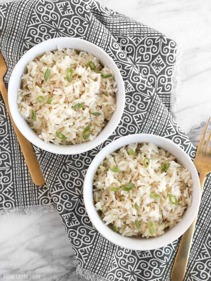 Overhead view of two bowls of cumin rice, garnished with green onion, sitting on a patterned napkin, two gold forks on the sides.