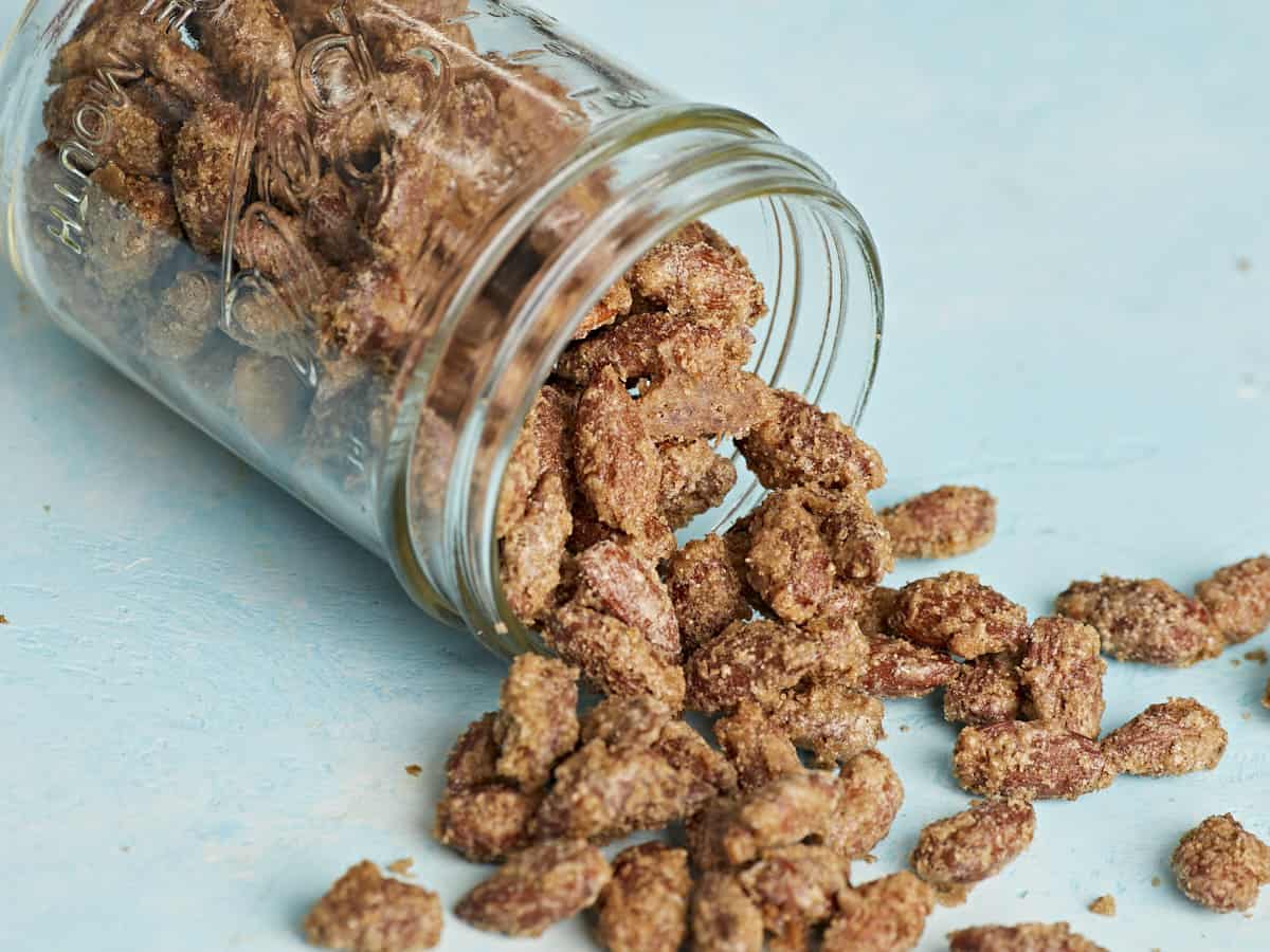 Side view of a mason jar full of candied almonds spilling onto a blue surface.