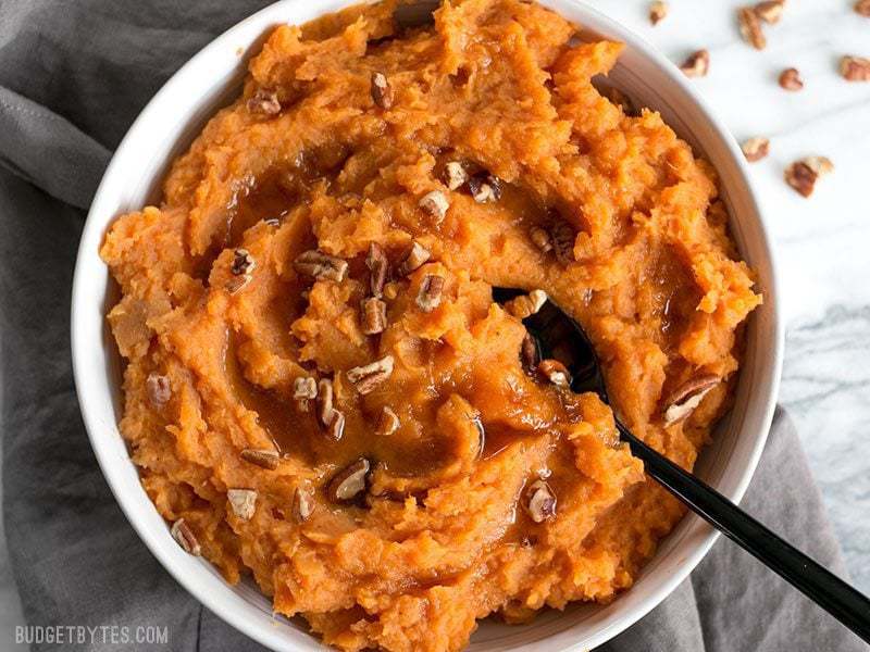 A spoon in a fluffy bowl of Maple Brown Butter Mashed Sweet Potatoes with pecans.