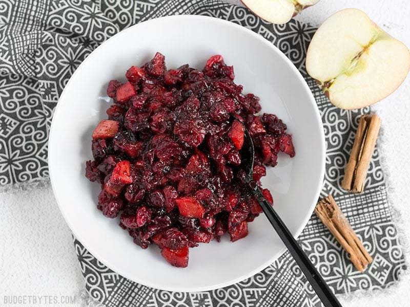 A bowl of thick Roasted Apple Cranberry Relish with a black spoon dug in, cinnamon sticks and apples on the side.