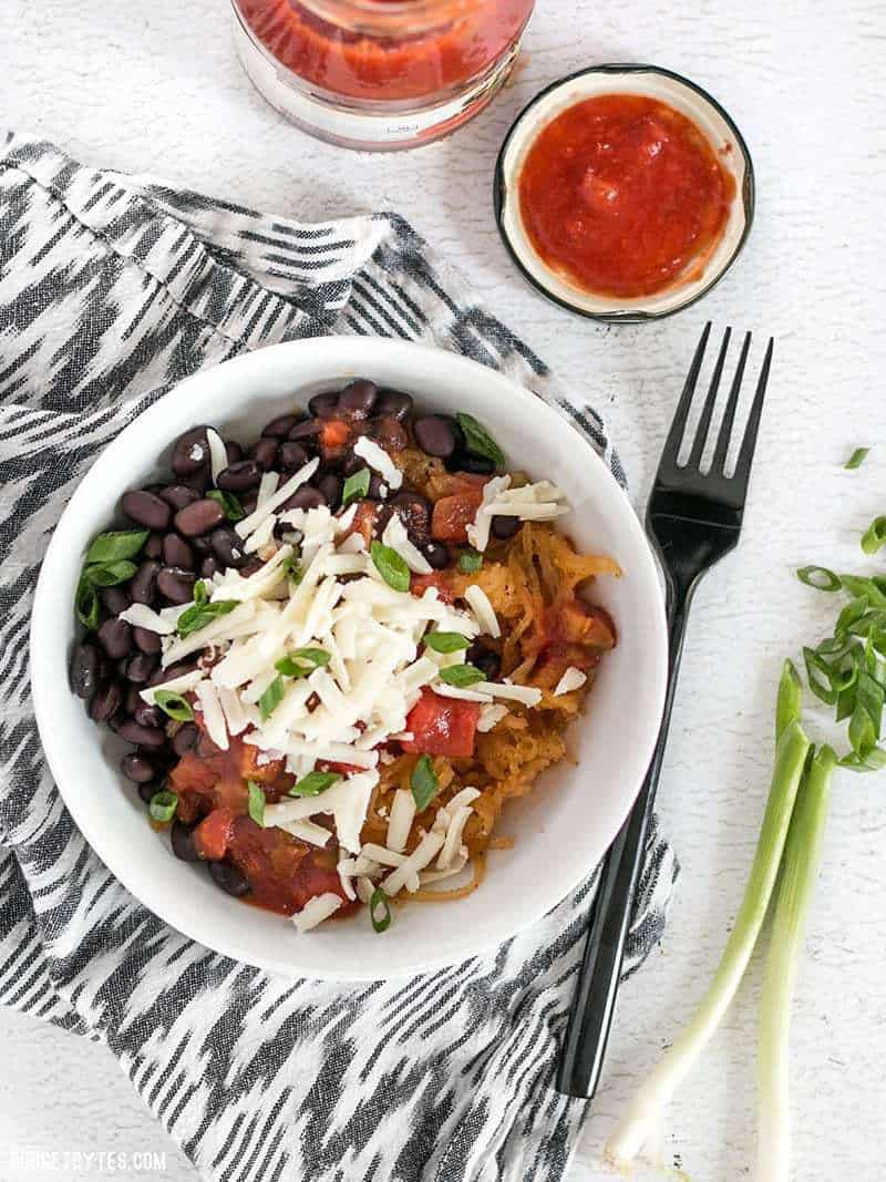 A southwest spaghetti squash bowl on a striped napkin with a jar of salsa nearby and a sliced green onion on the side.