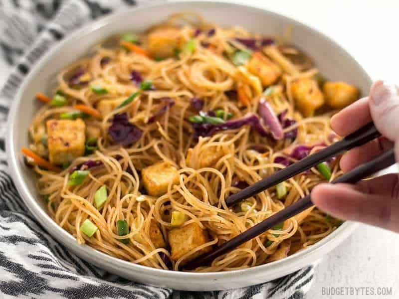 A bowl of Singapore Noodles with Crispy Tofu viewed from the front, chopsticks picking up a bunch of noodles