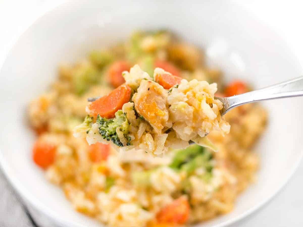 Close up of a forkful of cheesy chicken and rice casserole held in front of a bowl.