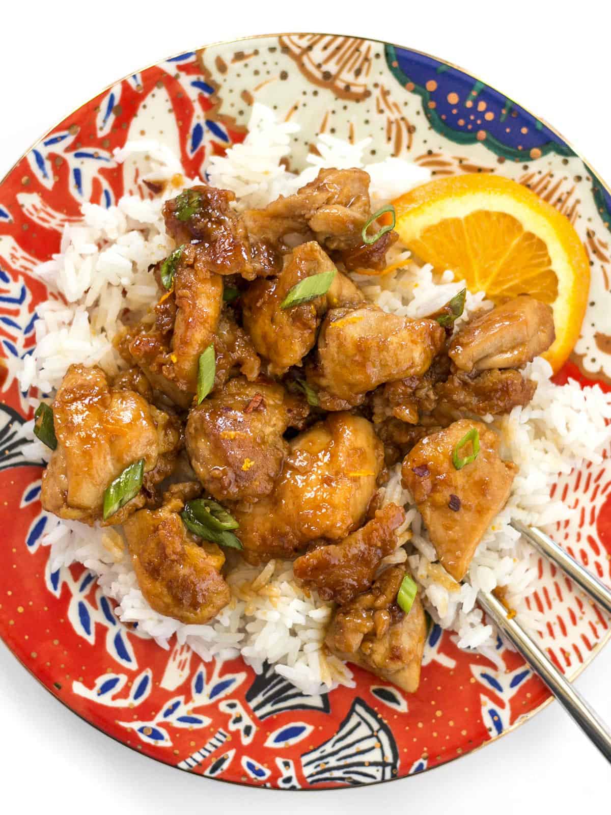 Overhead view of orange chicken on a plate with rice, green onion, and orange slices.