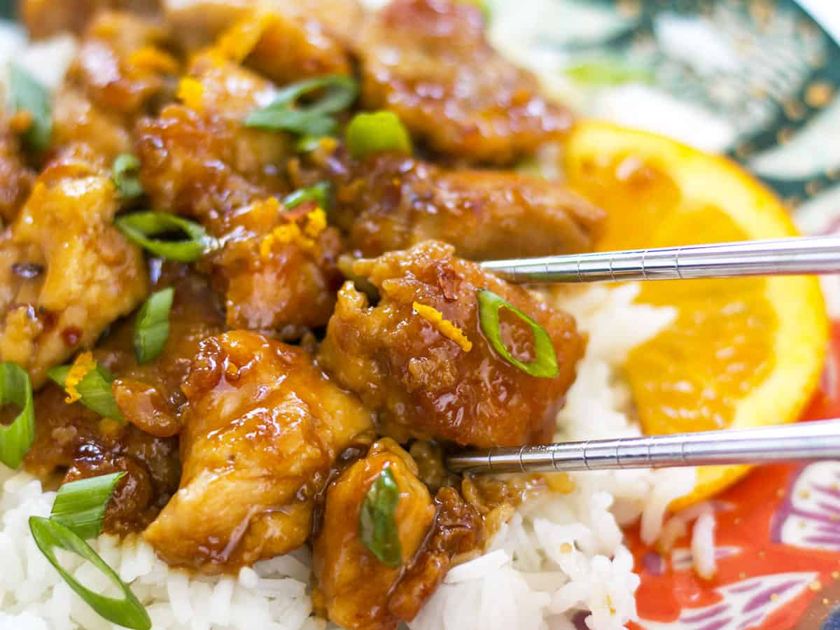 Close up of orange chicken on a plate with rice and chopsticks picking up one piece. 