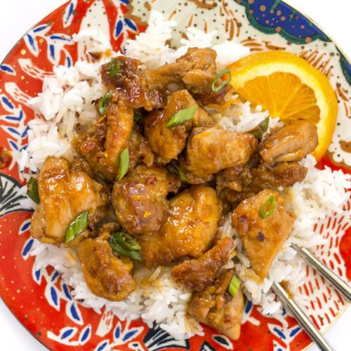 CLose up overhead view of orange chicken on a plate with rice, green onion, and orange slices.