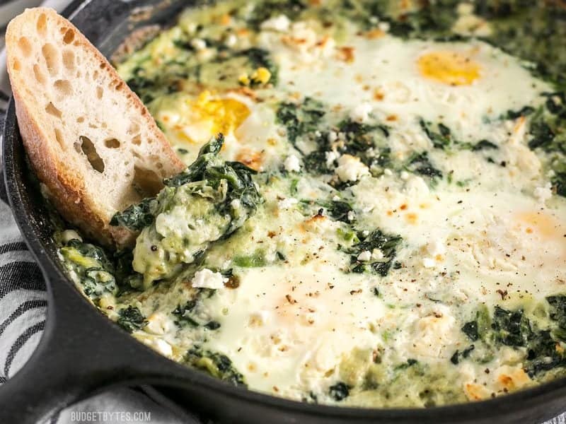 Close up shot of a piece of bread being dipped into Creamed Spinach Baked Eggs 