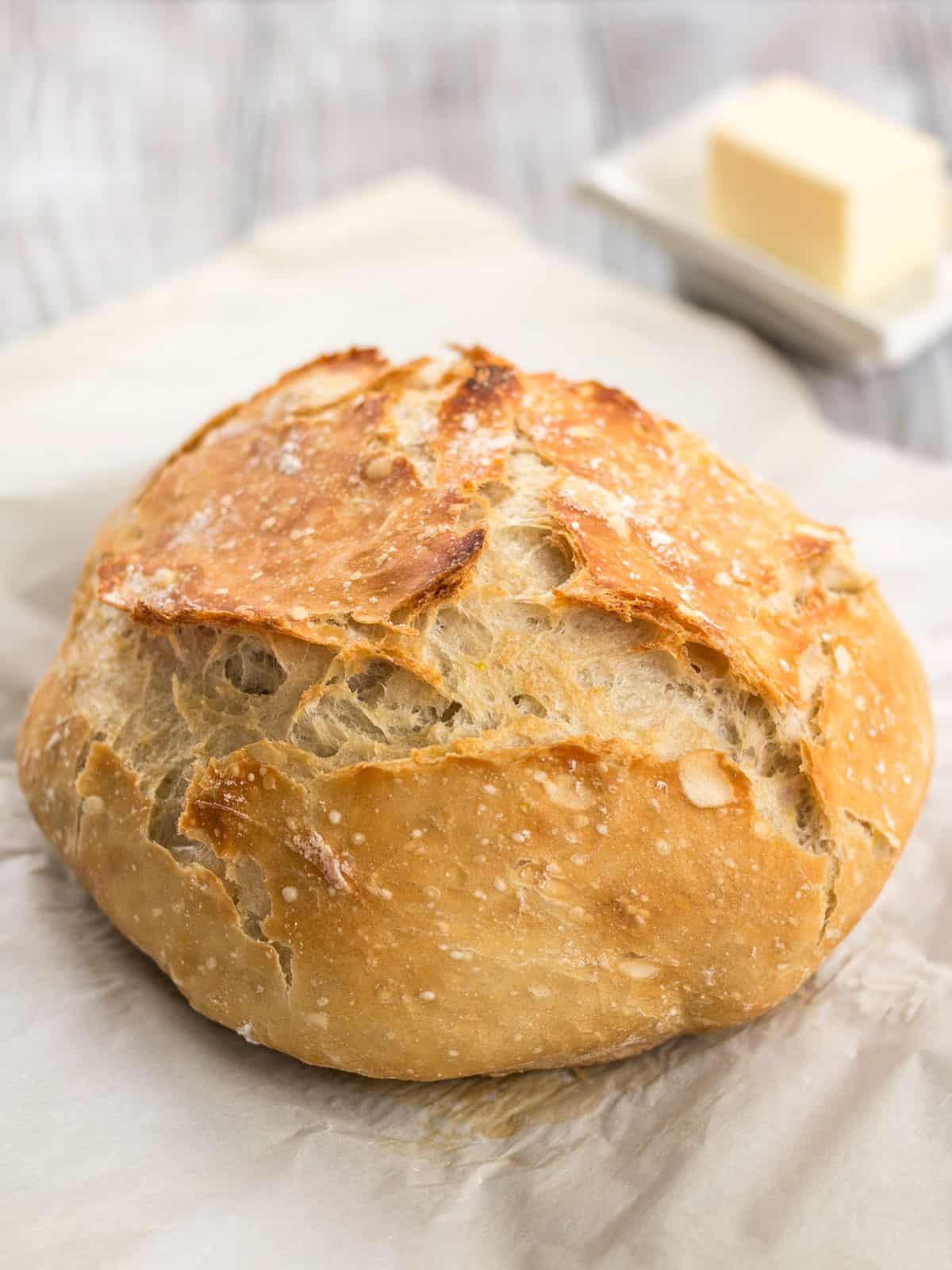 Side view of a baked loaf of no knead bread on parchment with butter in the background.