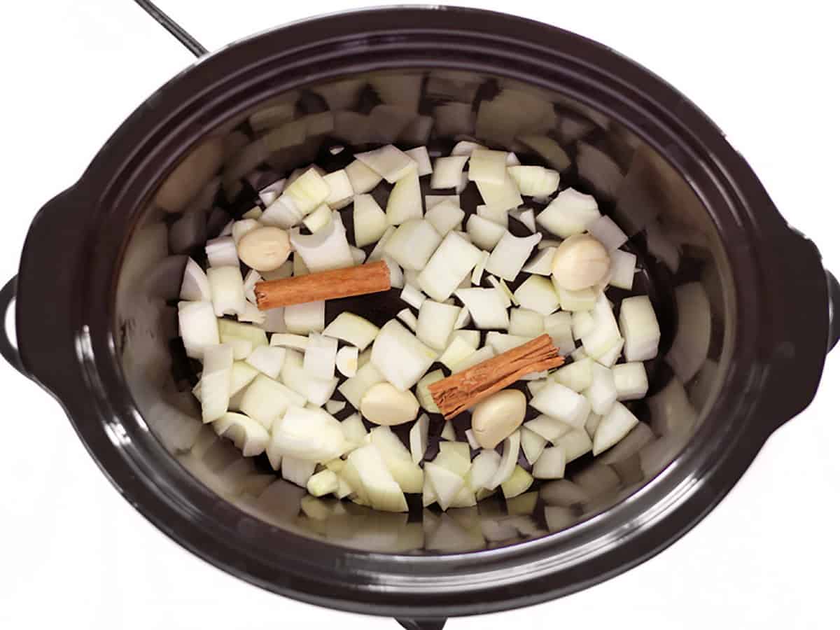 Diced onion, garlic, and cinnamon sticks in the bottom of a slow cooker.