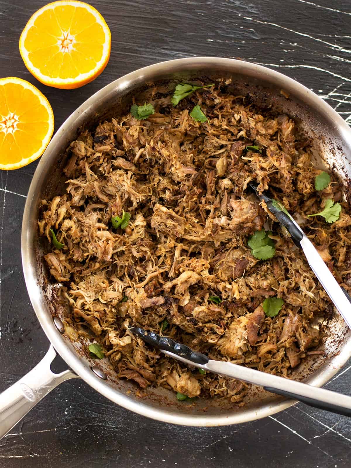 Overhead view of slow cooker carnitas in a skillet with tongs and an orange on the side.