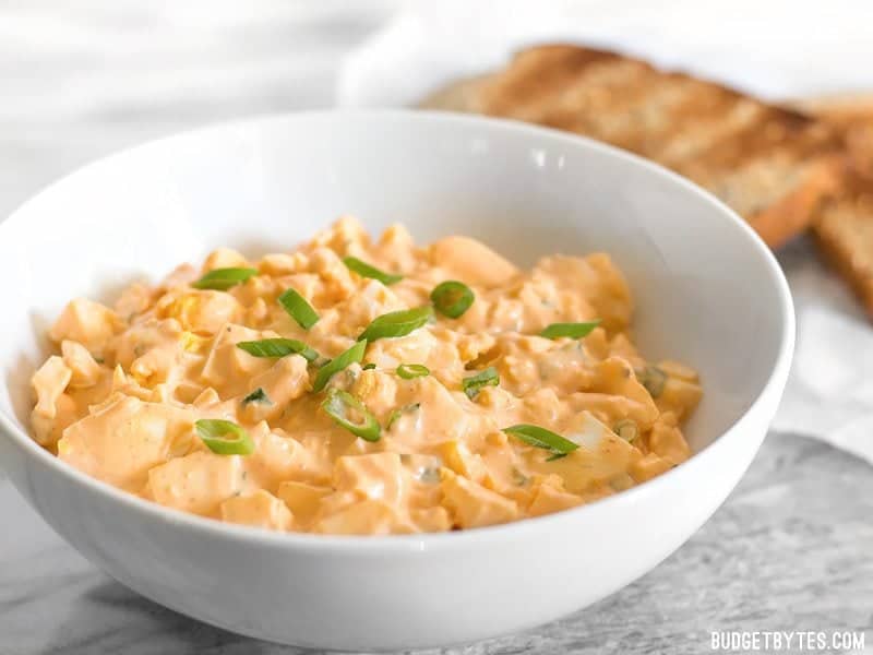 A bowl of Sriracha Egg Salad viewed from the side, toast in the background