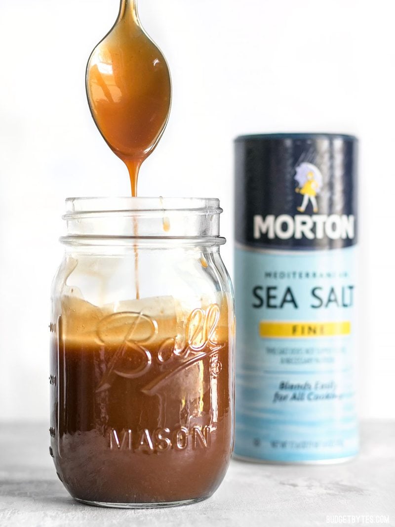 Homemade caramel dripping off a spoon into a mason jar with salt container in the background