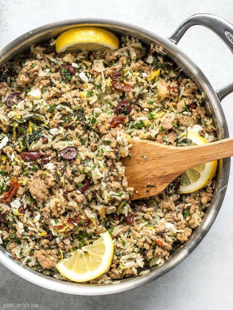 Overhead view of Greek Turkey and Rice Skillet in the skillet with a wooden spoon and lemon wedges.