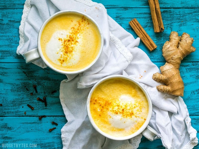 Two mugs of Golden Chai with cinnamon sticks and a ginger root on the side