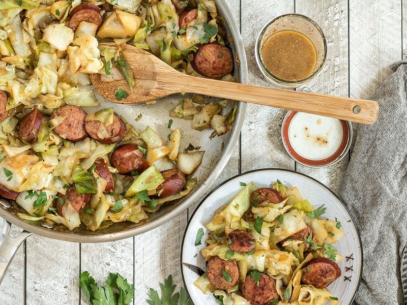 Kielbasa and Cabbage Skillet being served onto a plate from the skillet, with mustard vinaigrette in a jar on the side. 