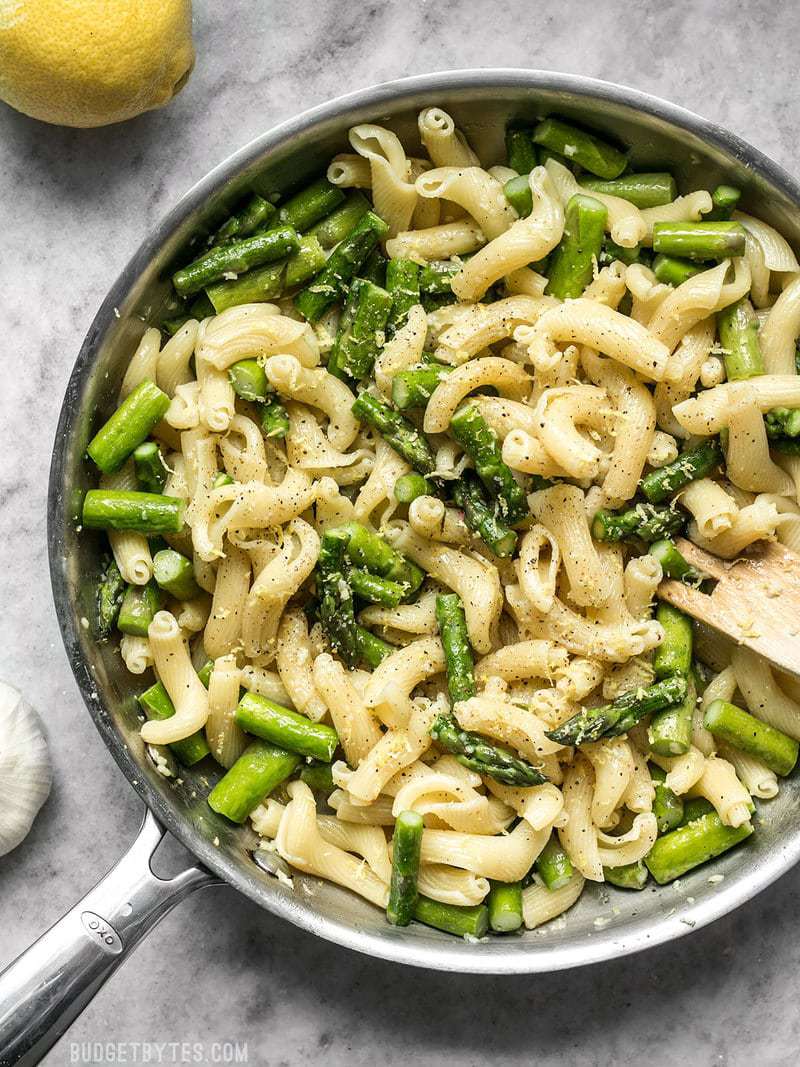 A skillet full of Lemon Garlic Asparagus Pasta with a wooden fork and lemons on the side