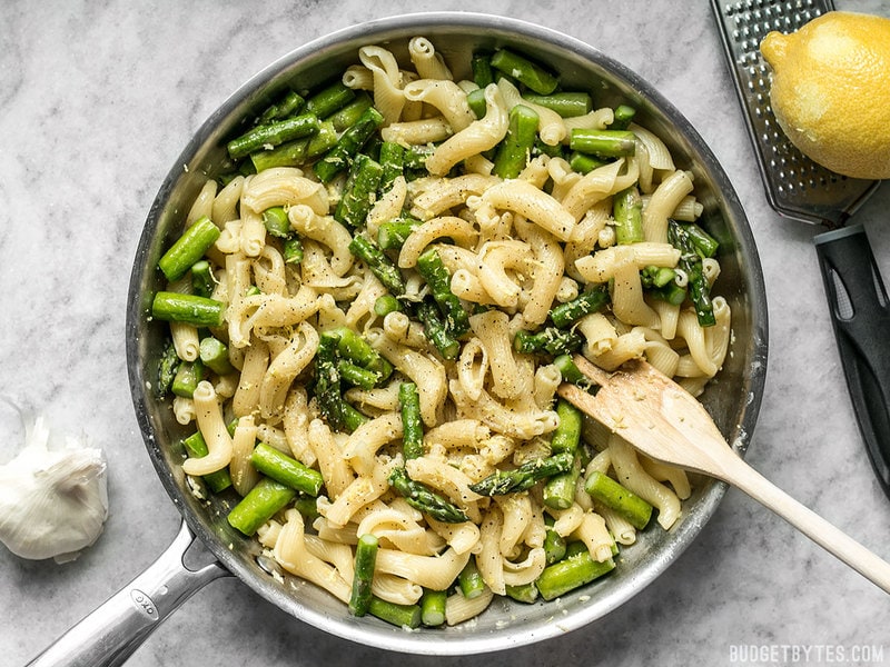 A skillet full of Lemon Garlic Asparagus Pasta with a wooden spoon and zested lemon on the side