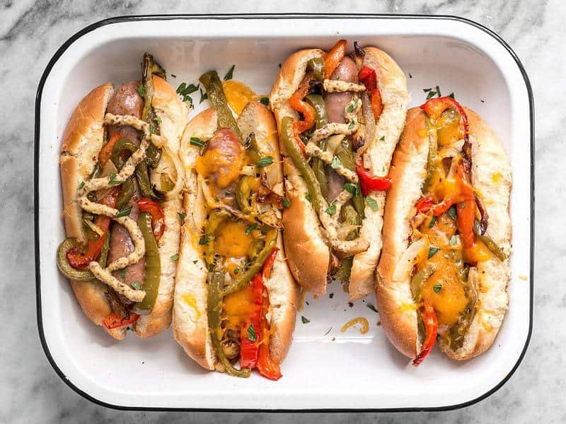 Four Roasted Bratwurst with Peppers and Onions, in buns with cheese and mustard, in a serving tray.