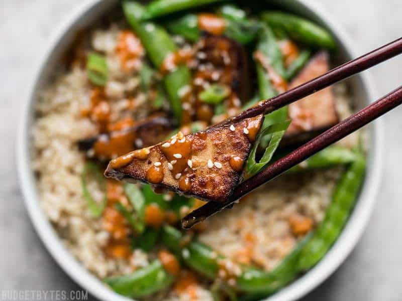 Chopsticks holding a piece of Soy Marinated Tofu with the bowl in the background