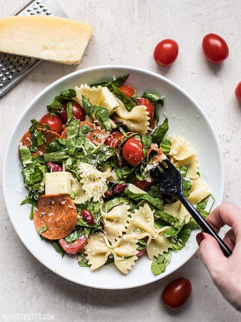 Pepperoni Pasta Salad being eaten with a black fork. Parmesan and tomatoes near by. 