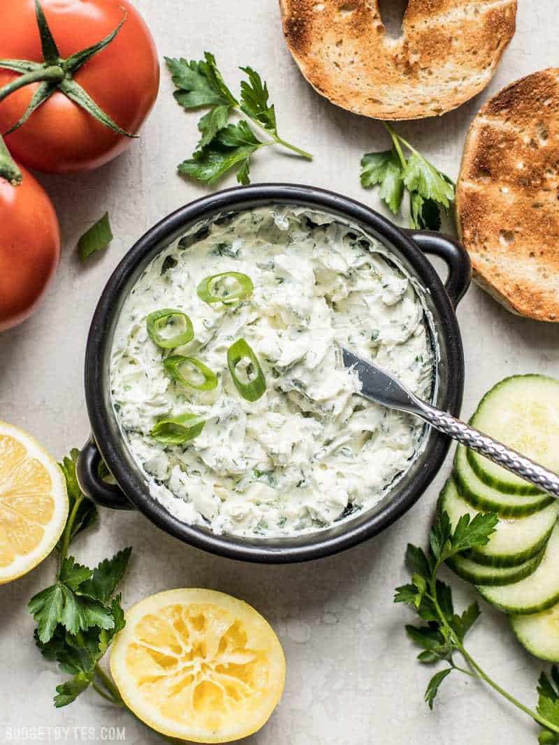 A bowl of Scallion Herb Cream Cheese Spread surrounded by vegetables, bagels, and lemons