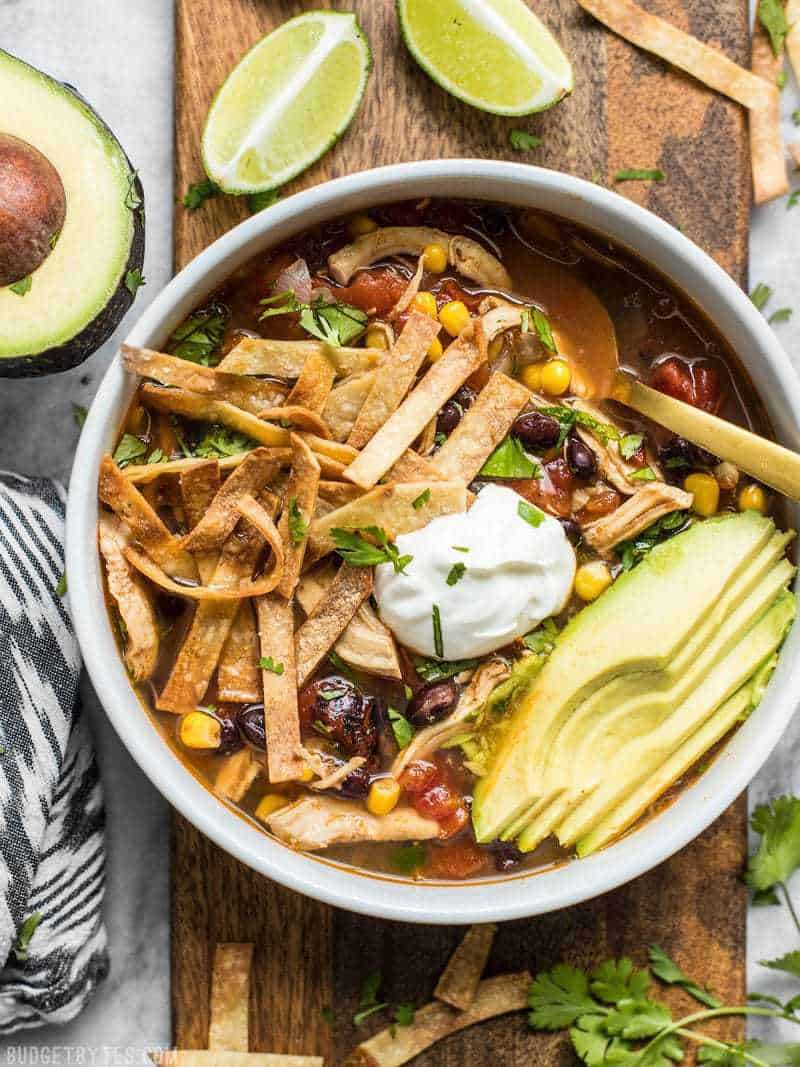 A bowl of Slow Cooker Chicken Tortilla Soup next to limes, avocado, and cilantro