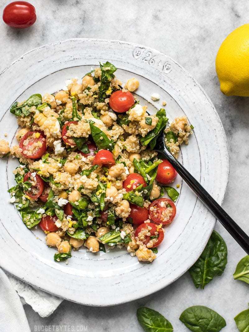 A plate full of Spinach Chickpea and Quinoa Salad with spinach, tomatoes, and lemons scattered around