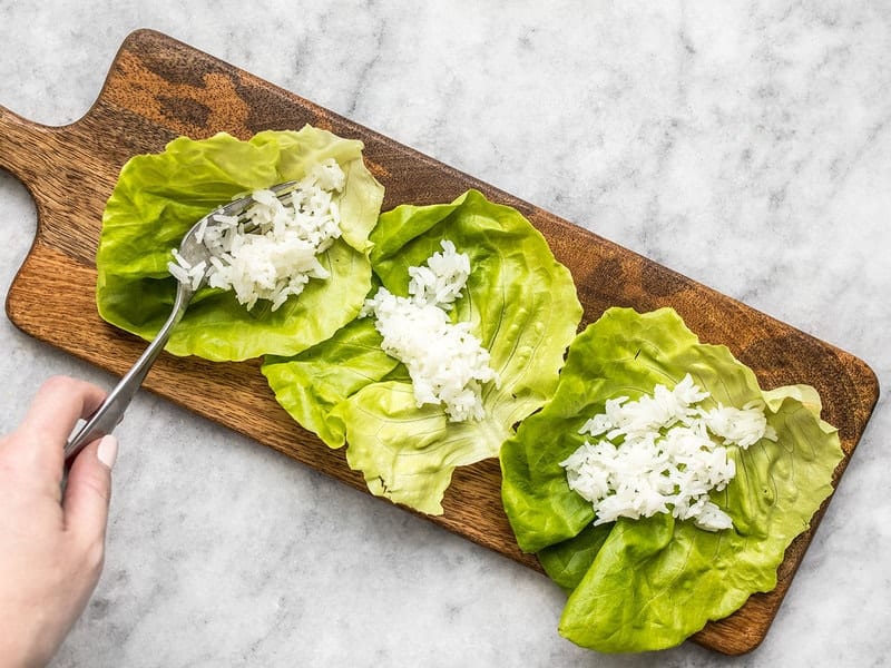 Rice being added to lettuce leaves on a wooden cutting board