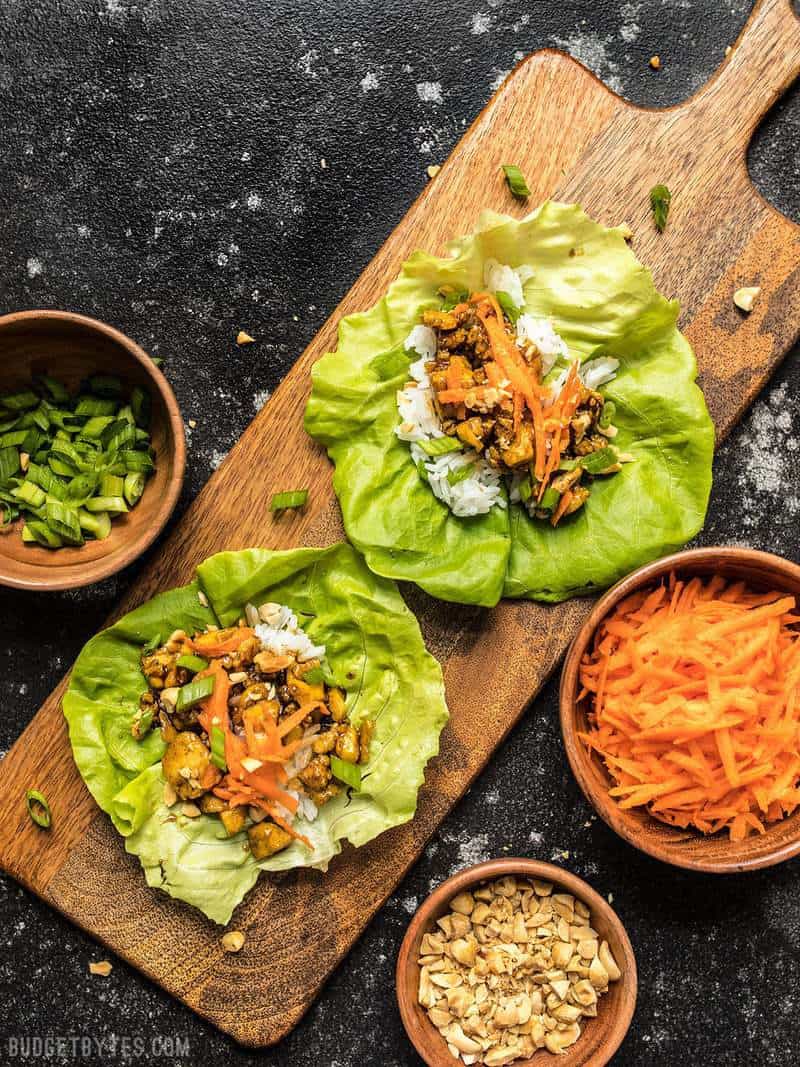 Two Hoisin Tofu Lettuce Cups on a wooden cutting board with bowls of toppings on the sides