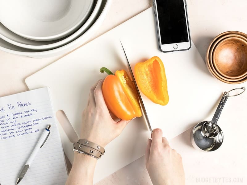 Hands cutting a yellow bell pepper with notes, bowls, measuring spoons, and a phone near by.