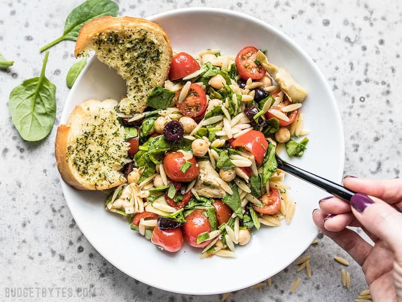 A hand taking a forkful of Italian Orzo Salad from a plate. Garlic Bread on the side. 