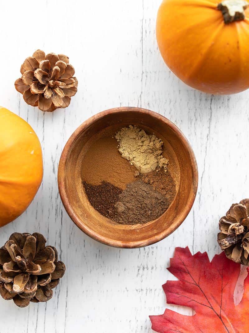 A small wooden bowl with spices, surrounded by pinecones, pumpkins, and leaves