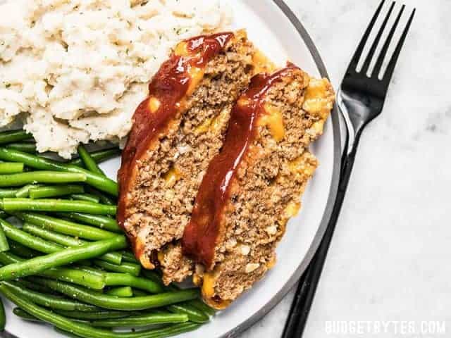 Two slices of Cheddar Cheeseburger Meatloaf on a dinner plate with mashed potatoes and green beans