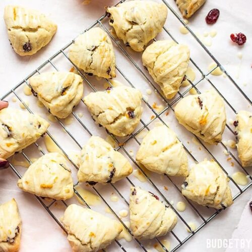 A hand taking one Cranberry Orange Cream Scone from the batch on a cooling rack.