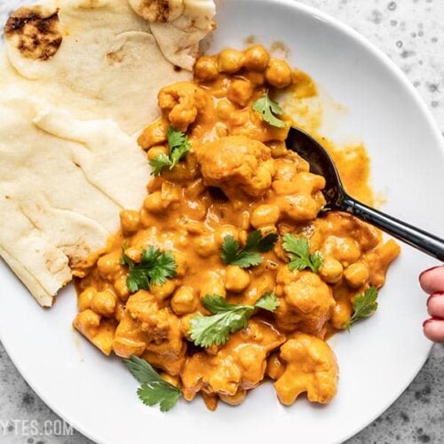 A bowl of creamy Cauliflower and Chickpea Masala being eaten with a spoon and two pieces of naan on the side.